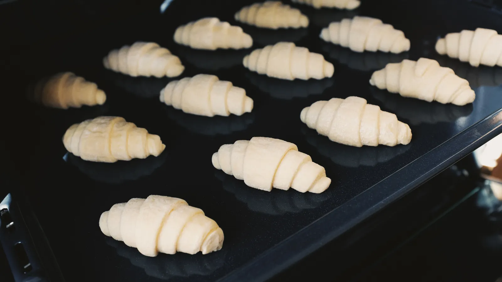 Ungebackene Croissants auf einem schwarzen Backblech im geöffneten Backofen